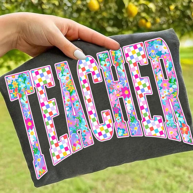 May include: Dark gray t-shirt with the word "TEACHER" in large, colorful letters. The letters are filled with a floral pattern and checkered accents, outlined in white. The shirt is held up by a hand.