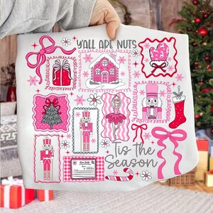 May include: A white Christmas-themed throw blanket with pink illustrations and text. The design includes a gingerbread house, nutcrackers, a Christmas tree, and the phrases "Y'all Are Nuts" and "Tis the Season".
