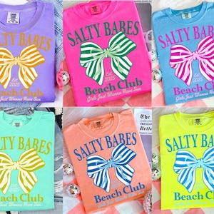 Może przedstawiać: Kolekcja sześciu kolorowych koszulek. Każda koszulka zawiera napis "SALTY BABES" nad grafiką kokardy i "Beach Club" poniżej. Koszulki są w kolorach fioletowym, r&oacute;żowym, niebieskim, zielonym, pomarańczowym i ż&oacute;łtym.