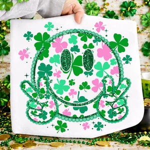May include: White St. Patrick's Day towel featuring a smiley face design with green and pink shamrocks. The smiley face has green glitter eyes and peace sign hands. The towel is decorated with a green lace border and shamrocks.
