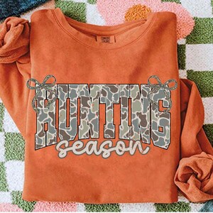May include: An orange long-sleeved sweatshirt with the words "HUNTING SEASON" in a camouflage pattern across the front. The word "HUNTING" is in large block letters, and "season" is in a script font. Decorative bows are on either side of the word "HUNTING".
