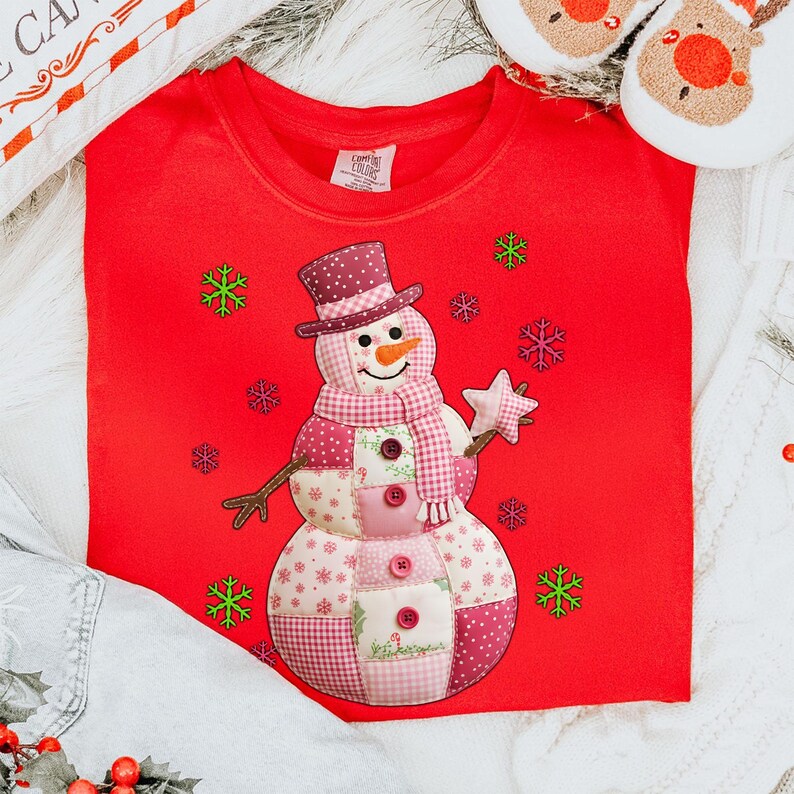 May include: A red sweatshirt with a patchwork snowman design. The snowman has a top hat, scarf, and star, and is decorated with buttons. Green and purple snowflakes surround the snowman. A festive holiday garment.