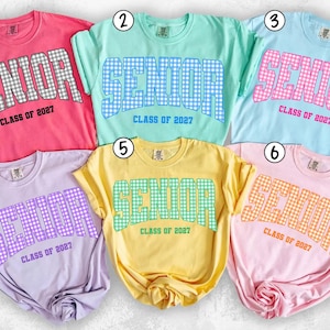 May include: Six short-sleeved crewneck t-shirts in pink, mint green, light blue, lavender, yellow, and light pink. Each shirt features the word SENIOR in a varsity font with a gingham pattern and the text CLASS OF 2027 printed underneath.