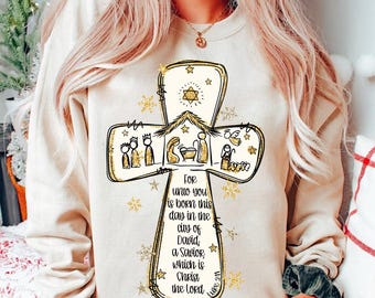 May include: A white sweatshirt with a gold glitter cross design featuring a nativity scene and the text "FOR unto you is born this day in the city of David, a Savior which is Christ the lord".