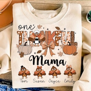 Può includere: Una felpa beige con il testo "one thankful mama" e un design a tema del Ringraziamento. Il design include un tacchino, zucche e foglie autunnali. La felpa presenta anche i nomi "Tom", "Susan", "Jayce" e "Emily" in un carattere più piccolo.