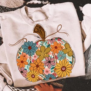 May include: Cream-coloured sweatshirt with a floral pumpkin design. The pumpkin is filled with colourful flowers in shades of blue, orange, yellow, and pink. The design includes a brown stem and vines, perfect for autumn.