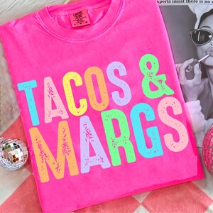 May include: Bright pink t-shirt with the words "TACOS & MARGS" in colorful, distressed lettering. The shirt is a Comfort Colors brand, and the design is centered on the front. A fun, casual top for taco and margarita lovers.