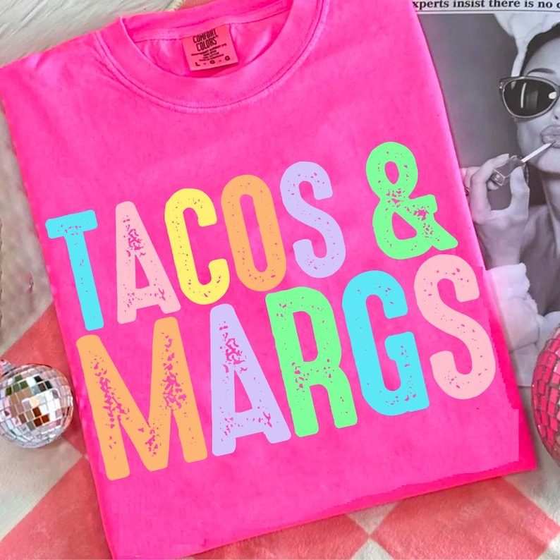 May include: Bright pink t-shirt with the words "TACOS & MARGS" in colorful, distressed lettering. The shirt is a Comfort Colors brand, and the design is centered on the front. A fun, casual top for taco and margarita lovers.