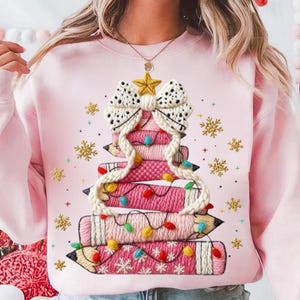 May include: A light pink sweatshirt featuring a Christmas tree design made of stacked pencils. The tree is adorned with a white bow, a gold star, and colorful string lights. Gold snowflakes are scattered around the tree.