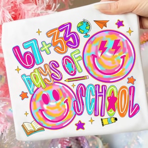 Może przedstawiać: Biała koszulka z kolorową grafiką świętującą '67 + 33 Days of School'. Projekt zawiera uśmiechnięte buzie, gwiazdki, globus, papierowy samolot i oł&oacute;wek, w jasnych kolorach.