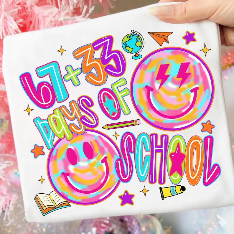 Może przedstawiać: Biała koszulka z kolorową grafiką świętującą '67 + 33 Days of School'. Projekt zawiera uśmiechnięte buzie, gwiazdki, globus, papierowy samolot i oł&oacute;wek, w jasnych kolorach.