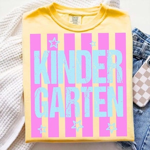 May include: Yellow t-shirt with the word "KINDERGARTEN" in light blue, set against pink vertical stripes. Small light blue stars are scattered around the text. The shirt is folded on a white surface with a pair of blue jeans and a phone.