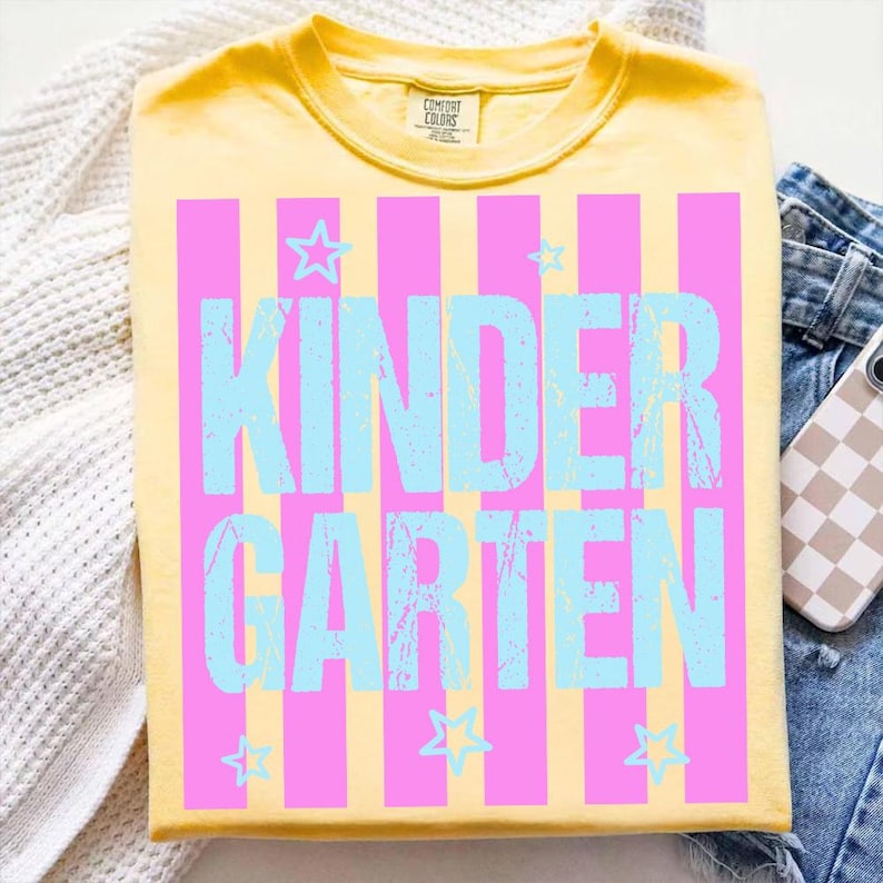 May include: Yellow t-shirt with the word "KINDERGARTEN" in light blue, set against pink vertical stripes. Small light blue stars are scattered around the text. The shirt is folded on a white surface with a pair of blue jeans and a phone.