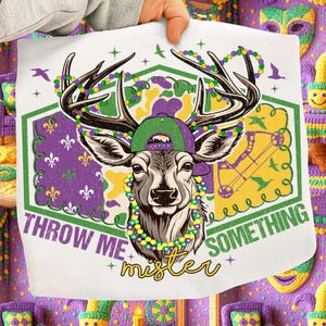 May include: White square fabric featuring a deer graphic wearing a green and purple cap and a beaded necklace. The text "THROW ME MISTER SOMETHING" is printed on the fabric. The deer has antlers and is surrounded by a green and purple hexagon.