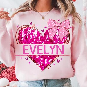 May include: Pink sweatshirt with a sequined heart and a pink bow. The heart has the name "EVELYN" written across it.