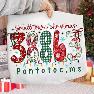 Puede incluir: Funda de almohada blanca con temática navideña con el texto "Small town christmas" y los números "3863" en patrones festivos. La palabra "Pontotoc, ms" está impresa debajo. La funda de almohada está decorada con copos de nieve y luces de cadena.