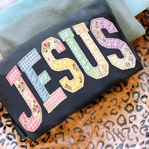 May include: A dark grey t-shirt with the word "JESUS" appliquéd in colourful, patchwork-style letters. Each letter features a different pattern, including floral prints, polka dots, and gingham, with a stitched border.