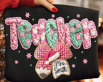 May include: Black sweatshirt with the word "TEACHER" embroidered in pink and green, with a bow and Santa hat design. The name "Mrs. Smith" is written below the bow. Pink stars are scattered around the design.