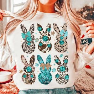 May include: White sweatshirt with a graphic print of six Easter bunnies. The bunnies are wearing bows and have a variety of animal print patterns.