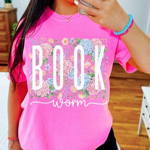 May include: A vibrant pink t-shirt featuring the text "BOOK worm" in white, framed by a floral arrangement in pastel hues. The design incorporates blue hydrangeas, pink roses, and lavender. The shirt is a relaxed fit.