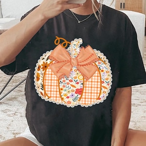 K&ouml;nnte beinhalten: Schwarzes T-Shirt mit einem K&uuml;rbis-Design. Der K&uuml;rbis besteht aus floralen, karierten und Spitzenmustern mit einer orangefarbenen Schleife. Das Design ist auf dem T-Shirt zentriert und schafft ein festliches Herbstthema.