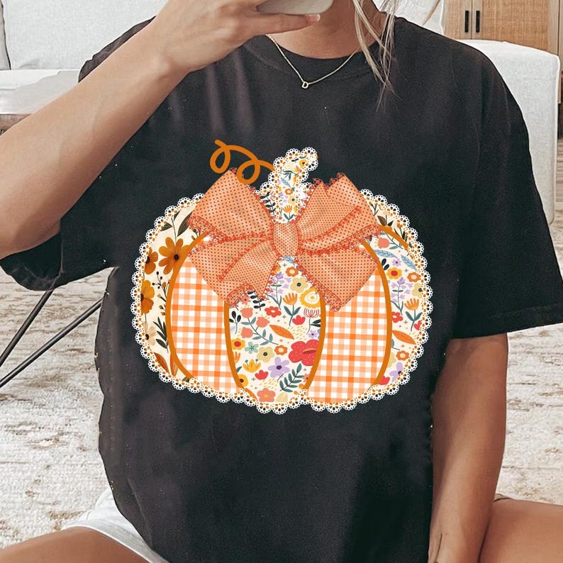 K&ouml;nnte beinhalten: Schwarzes T-Shirt mit einem K&uuml;rbis-Design. Der K&uuml;rbis besteht aus floralen, karierten und Spitzenmustern mit einer orangefarbenen Schleife. Das Design ist auf dem T-Shirt zentriert und schafft ein festliches Herbstthema.