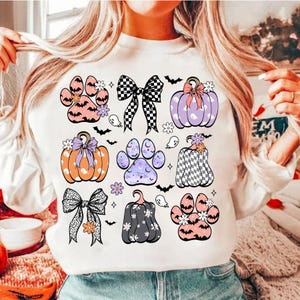 Puede incluir: Sudadera blanca con un diseño de temática de Halloween con calabazas, huellas de patas, lazos y murciélagos. Las calabazas están decoradas con lunares, cuadros y flores. Las huellas de patas son moradas, rosas y azules con detalles de purpurina.