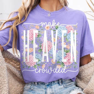 May include: A lilac-colored t-shirt with the words "make HEAVEN crowded" in white. The word "HEAVEN" is filled with pink and blue hydrangea flowers. The shirt has a crew neck and short sleeves.