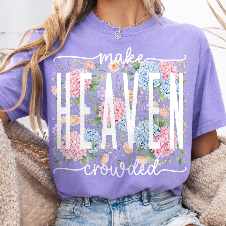 May include: A lilac-colored t-shirt with the words "make HEAVEN crowded" in white. The word "HEAVEN" is filled with pink and blue hydrangea flowers. The shirt has a crew neck and short sleeves.
