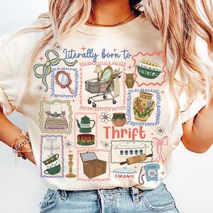 May include: Cream-colored sweatshirt with a colorful graphic design featuring the text "Literally born to Thrift." The design includes illustrations of vintage items like a shopping cart, owl, teapot, and telephone.