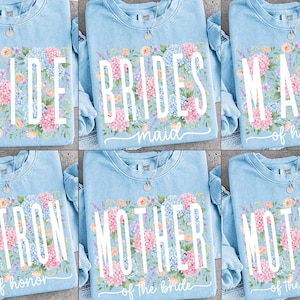 May include: Light blue crewneck sweatshirts with floral designs and text for a bridal party. The text includes "Bride," "Bridesmaid," "Maid of Honor," "Matron of Honor," "Mother of the Bride," and "Mother of the Groom."