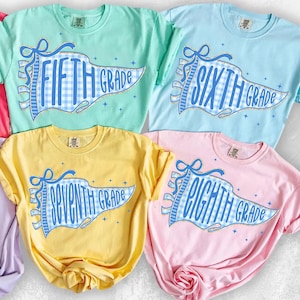 May include: Four crew neck t-shirts in mint green, light blue, yellow, and light pink. Each shirt features a blue and white gingham pennant graphic with gold glitter outlines. The text reads FIFTH GRADE, SIXTH GRADE, SEVENTH GRADE, and EIGHTH GRADE.