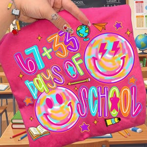 Może przedstawiać: Jasnor&oacute;żowa koszulka z napisem "67 + 35 Days of School" w kolorowych literach. Projekt zawiera uśmiechnięte buzie, gwiazdki, globus i papierowy samolot. Koszulka jest prawdopodobnie dla uczni&oacute;w.