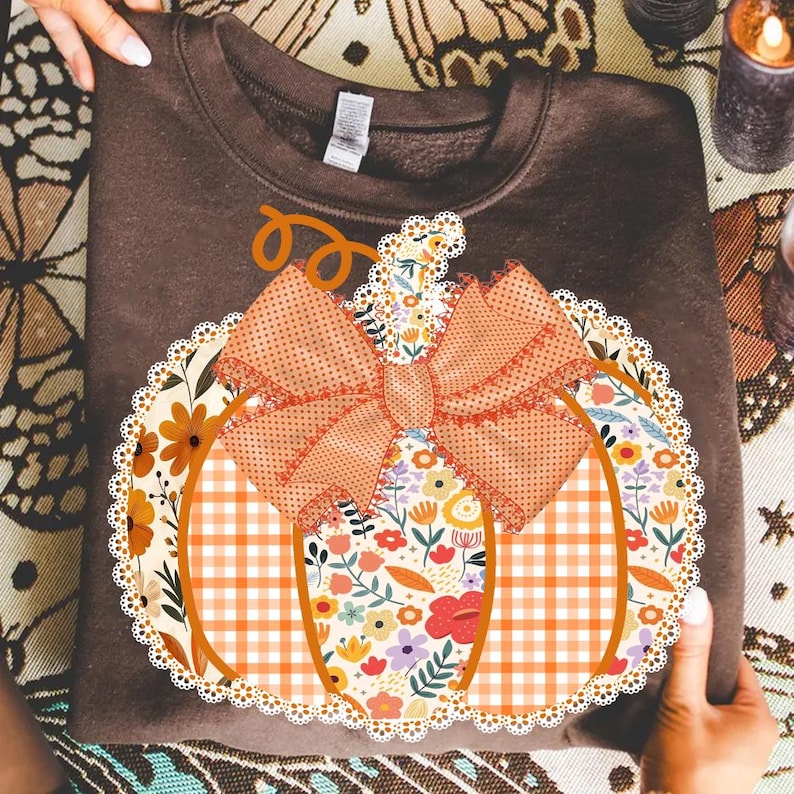 K&ouml;nnte beinhalten: Ein braunes Sweatshirt mit einem K&uuml;rbisdesign. Der K&uuml;rbis ist mit einem Blumenmuster, orangefarbenem Vichy-Karo und einer gro&szlig;en Schleife verziert. Das Design ist mit einem wei&szlig;en Spitzensaum versehen, ideal f&uuml;r den Herbst.
