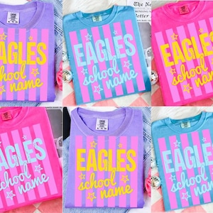 May include: A collection of six t-shirts in various colors, including purple, blue, and pink. Each shirt features the word "EAGLES" in large letters, along with "school name" and star graphics, all set against a striped background.