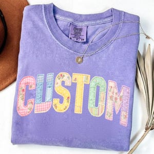 May include: A lavender Comfort Colors t-shirt with the word "CUSTOM" appliqued across the front in a patchwork design. The letters are in various floral and patterned fabrics. A silver necklace with a pendant is draped across the shirt.