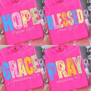 May include: Four hot pink t-shirts with inspirational words in colorful block letters. The words are "HOPE", "BLESSED", "GRACE", and "PRAY". Each shirt has a corresponding Bible verse printed below the word.
