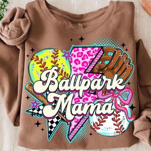 Puede incluir: Sudadera marr&oacute;n con un dise&ntilde;o gr&aacute;fico colorido. El dise&ntilde;o incluye una pelota de b&eacute;isbol, un guante de b&eacute;isbol, un rayo con estampado de leopardo rosa y el texto "Ballpark Mama". La sudadera tiene mangas largas.