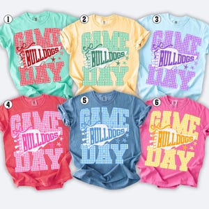 以下が含まれることがあります： ミント、イエロー、ライトブルー、レッド、デニムブルー、ピンクの半袖Tシャツ6枚。各シャツには、ギンガムチェック柄のグラフィックがプリントされており、ペナントに「GAME DAY」と「BULLDOGS」の文字、そして同系色の小さな星のアイコンが描かれています。