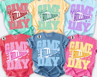 Könnte beinhalten: Sechs kurzärmelige T-Shirts in Mint, Gelb, Hellblau, Rot, Denimblau und Pink. Jedes Shirt zeigt eine Grafik mit Vichy-Karo-Muster, dem Text GAME DAY und BULLDOGS auf einem Wimpel sowie kleinen Sternsymbolen in den jeweils passenden Farben.