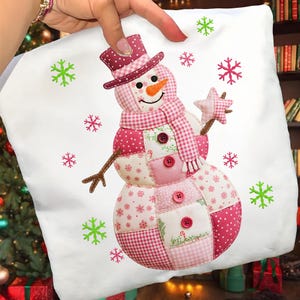 May include: A white cushion featuring a patchwork snowman design. The snowman has a pink top hat, scarf, and buttons, with a carrot nose and a star-shaped wand. Green and pink snowflakes are scattered around the snowman.