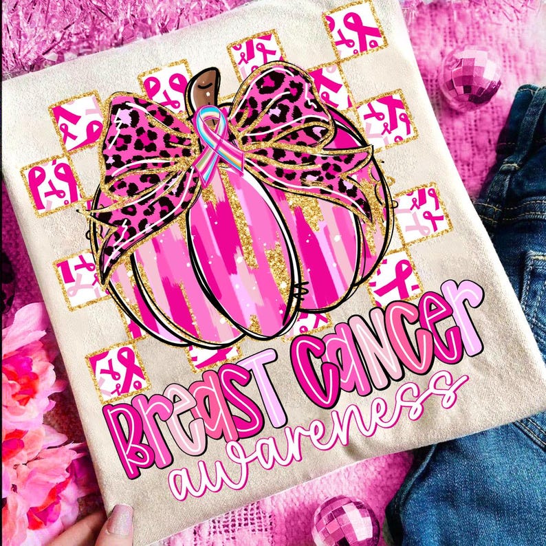 May include: A beige t-shirt with a pink pumpkin design and the words "Breast Cancer Awareness". The pumpkin has a pink and gold color scheme with a leopard print bow and a pink ribbon.