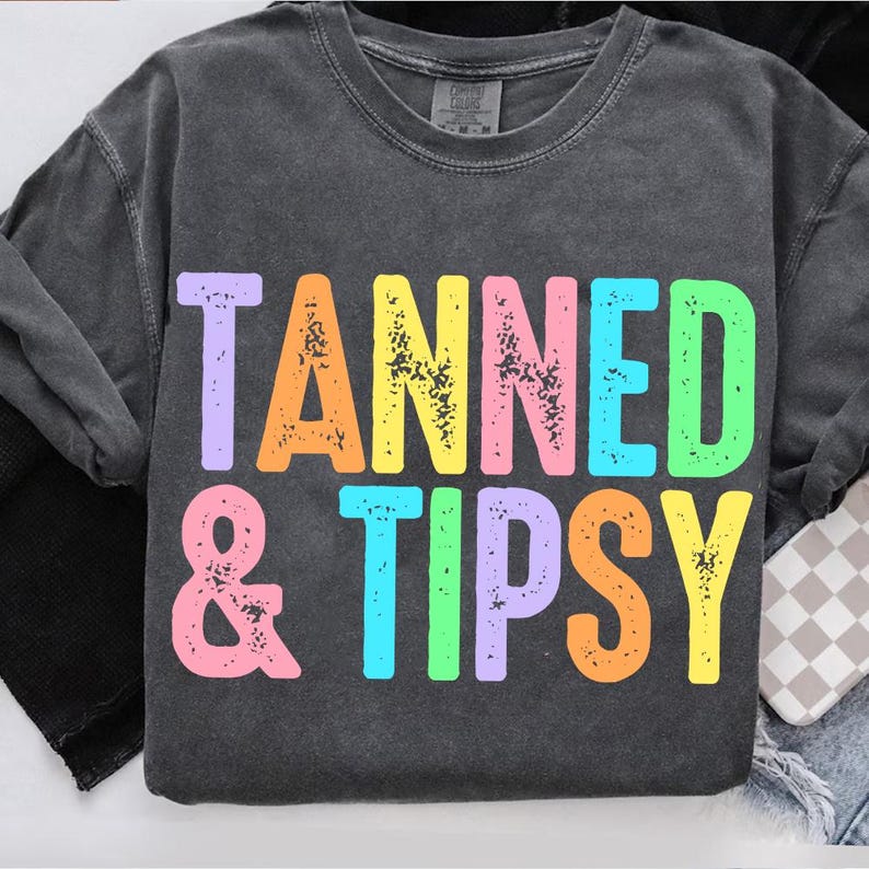 May include: Dark gray sweatshirt with the words "TANNED & TIPSY" in colorful, block letters. The text is in shades of purple, orange, blue, green, and yellow. The sweatshirt has a crew neck and long sleeves.
