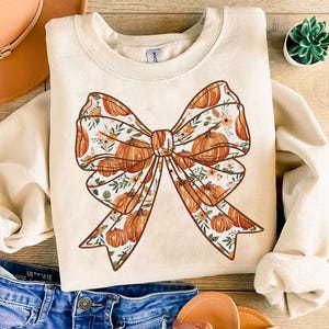 Op de afbeelding: Een crèmekleurige trui met een grote bruine strik die op de voorkant is gedrukt. De strik is versierd met een patroon van oranje pompoenen en witte bloemen.