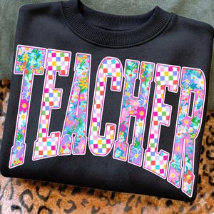 May include: Black sweatshirt with the word "TEACHER" in large, colorful letters. The letters are filled with a floral pattern and a checkered design, outlined in pink. The sweatshirt has a crew neck.