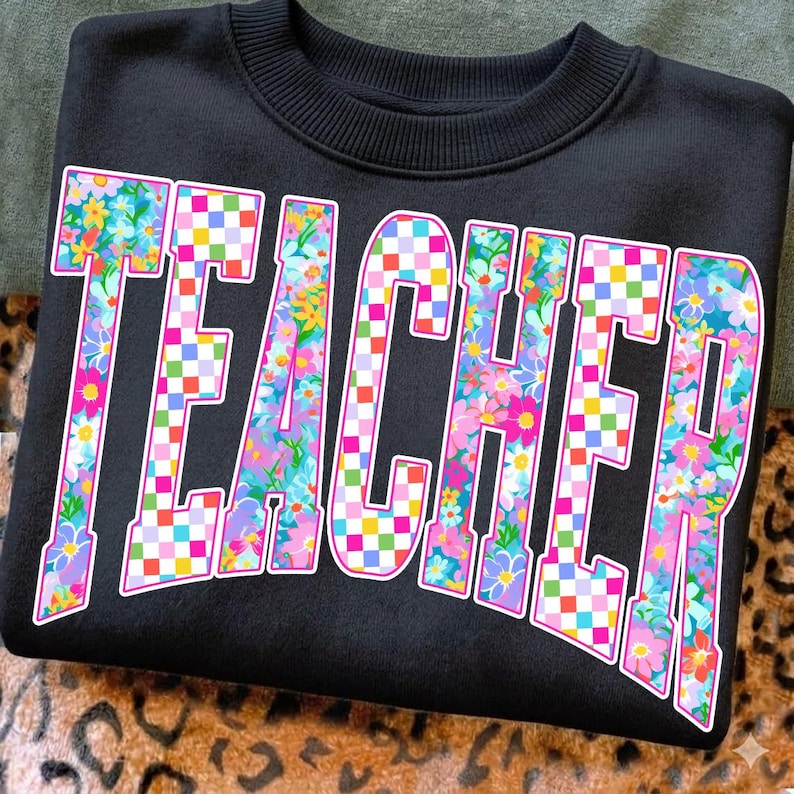 May include: Black sweatshirt with the word "TEACHER" in large, colorful letters. The letters are filled with a floral pattern and a checkered design, outlined in pink. The sweatshirt has a crew neck.