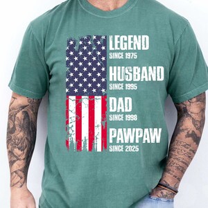 K&ouml;nnte beinhalten: Ein gr&uuml;nes T-Shirt mit einem verblassten Design der amerikanischen Flagge. Die Flagge ist vertikal ausgerichtet und zeigt den Text "LEGEND SINCE 1975", "HUSBAND SINCE 1995", "DAD SINCE 1998" und "PAWPAW SINCE 2025" in wei&szlig;en Blockbuchstaben.