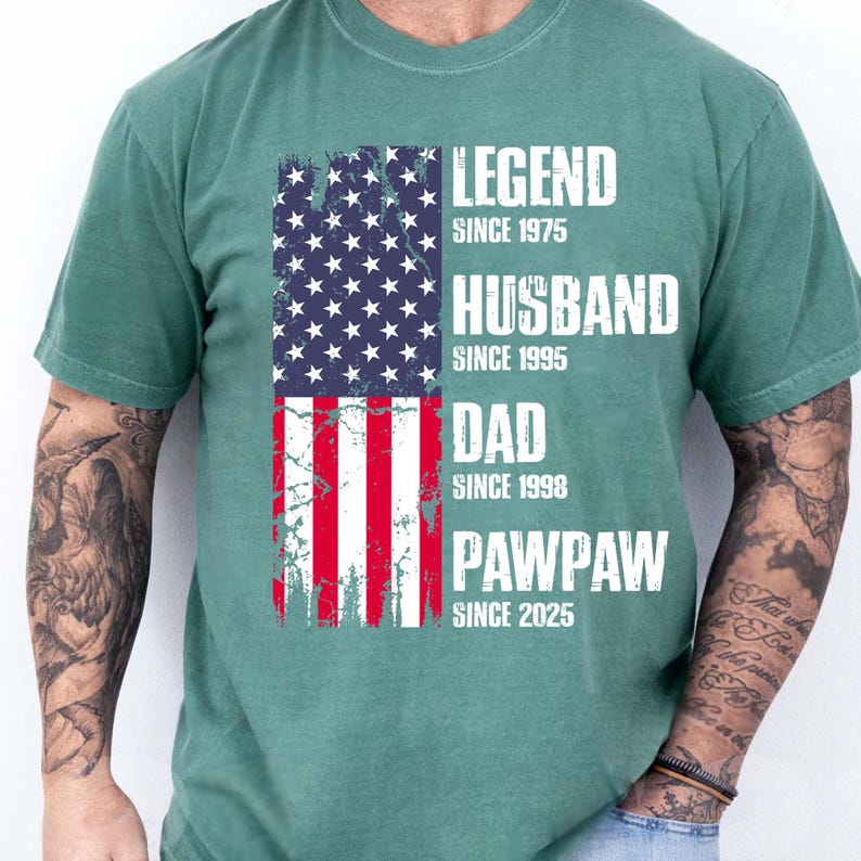 K&ouml;nnte beinhalten: Ein gr&uuml;nes T-Shirt mit einem verblassten Design der amerikanischen Flagge. Die Flagge ist vertikal ausgerichtet und zeigt den Text "LEGEND SINCE 1975", "HUSBAND SINCE 1995", "DAD SINCE 1998" und "PAWPAW SINCE 2025" in wei&szlig;en Blockbuchstaben.