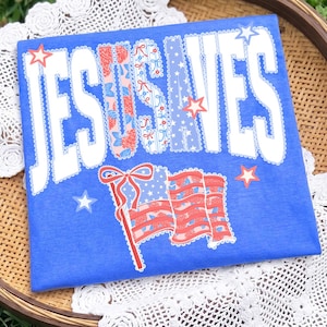May include: Blue t-shirt with the words "JESUS SAVES" in white, scalloped letters. The letter "S" is decorated with floral and bow patterns. An American flag graphic and star accents complete the patriotic design.