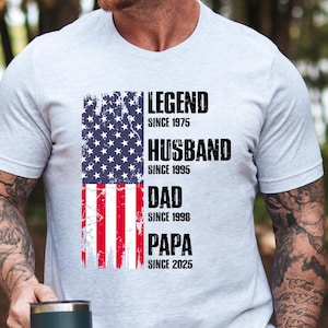 K&ouml;nnte beinhalten: Ein hellgraues T-Shirt mit einem verblassten amerikanischen Flaggen-Design. Das Shirt zeigt die Worte "LEGEND SINCE 1975", "HUSBAND SINCE 1995", "DAD SINCE 1998" und "PAPA SINCE 2025" in fetter schwarzer Schrift.